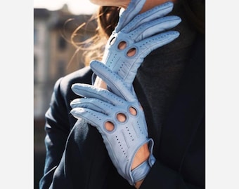 May include: A pair of light blue leather driving gloves featuring circular knuckle cutouts and perforated detailing along the fingers and back of the hand. The gloves have a wrist strap with a snap closure and a smooth, matte finish texture.
