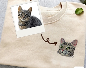 Puede incluir: Una sudadera beige con un gato marrón bordado en la parte delantera. El gato está mirando hacia la derecha. La sudadera está doblada por la mitad.