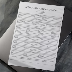 May include: A blank application form for employment, printed on white paper. The form includes fields for personal information, education, and previous employment history. The document is placed on a dark gray surface.