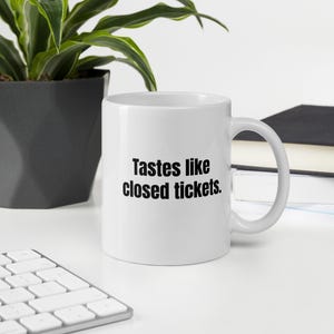 May include: White ceramic coffee mug with the text "Tastes like closed tickets" in black. The mug is on a white surface with a keyboard, a potted plant, and a stack of books in the background.