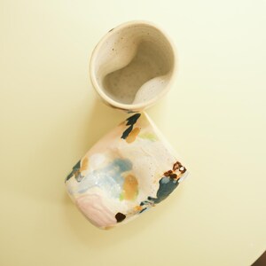 May include: Two ceramic mugs with a speckled, off-white finish. One mug is upright, the other is tilted, revealing a colorful, abstract design of pink, blue, and brown brushstrokes. The mugs are on a pale yellow surface.