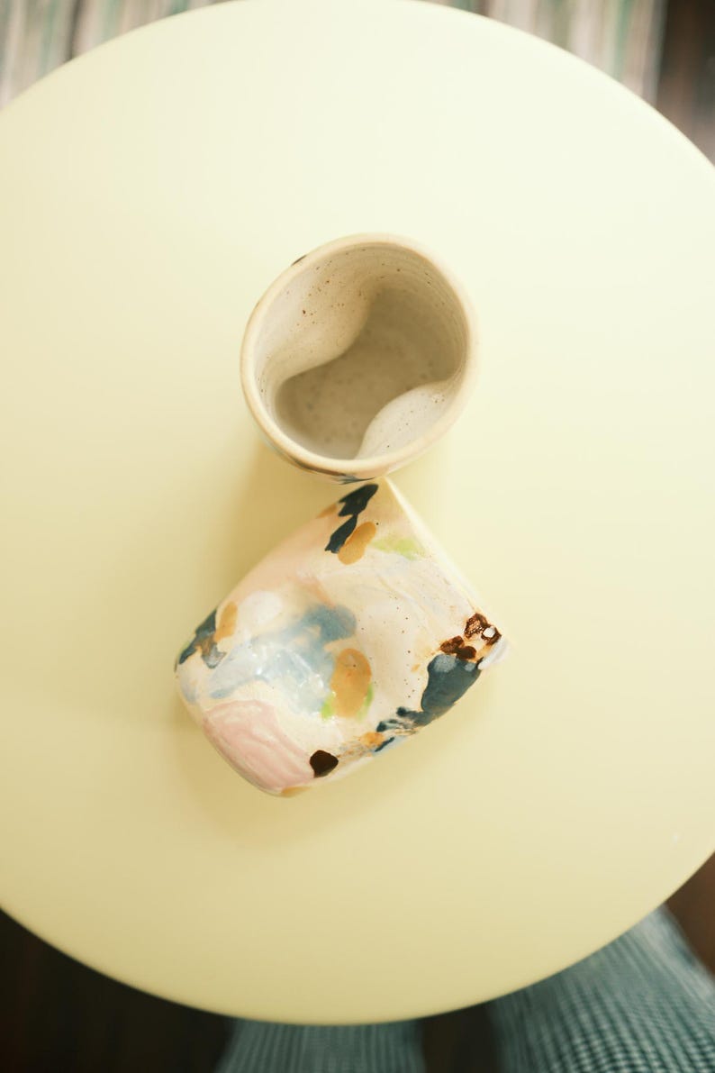 May include: Two ceramic mugs with a speckled, off-white finish. One mug is upright, the other is tilted, revealing a colorful, abstract design of pink, blue, and brown brushstrokes. The mugs are on a pale yellow surface.