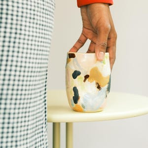 May include: A ceramic tumbler with an abstract design. The cup features a beige base with colorful brushstrokes in shades of blue, brown, and peach. The cup is held by a person wearing a red sweater.