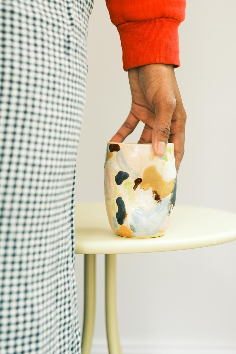 May include: A ceramic tumbler with an abstract design. The cup features a beige base with colorful brushstrokes in shades of blue, brown, and peach. The cup is held by a person wearing a red sweater.