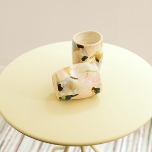 May include: Two ceramic cups with abstract painted designs in pastel colors, including pink, blue, and yellow, sit on a round, pale yellow table. The cups have a speckled, off-white base and are placed in front of a white wall.