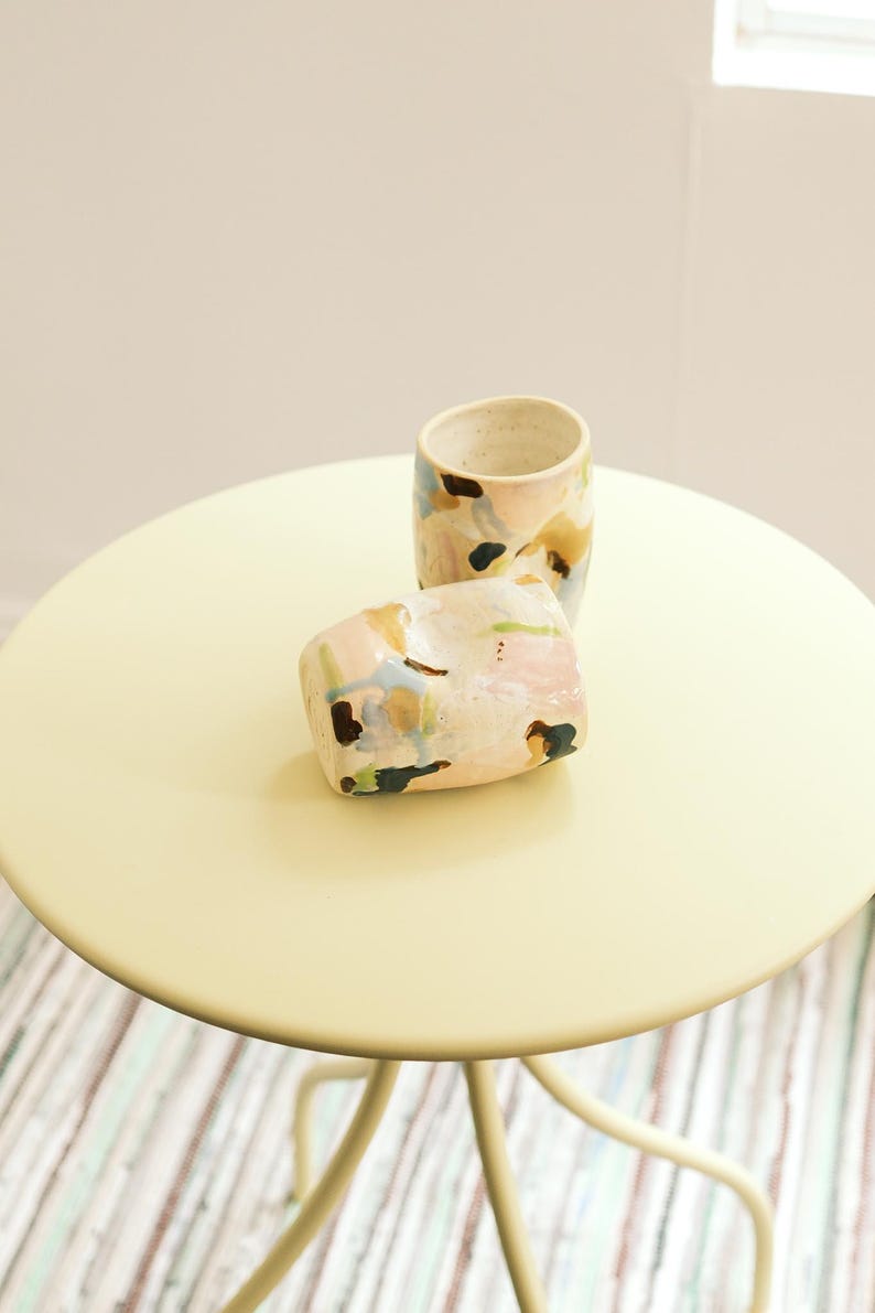 May include: Two ceramic cups with abstract painted designs in pastel colors, including pink, blue, and yellow, sit on a round, pale yellow table. The cups have a speckled, off-white base and are placed in front of a white wall.