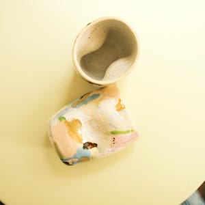 May include: Two handmade ceramic cups. One is a light beige color with a unique, curved interior. The other cup is a similar shape, but with a colorful, abstract design featuring pink, blue, and gold hues. The cups are on a yellow surface.