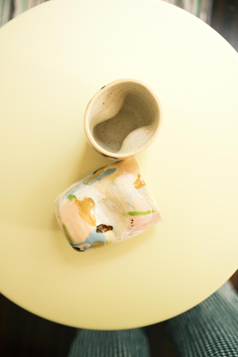 May include: Two handmade ceramic cups. One is a light beige color with a unique, curved interior. The other cup is a similar shape, but with a colorful, abstract design featuring pink, blue, and gold hues. The cups are on a yellow surface.