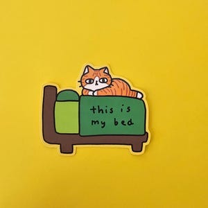 May include: A sticker showing a cartoon cat with orange and white stripes on a green bed with a brown frame. The text "this is my bed" is on the green blanket. The background is yellow.