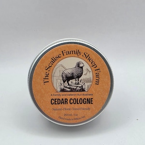 Op de afbeelding: Een ronde zilveren metalen blikje met ceder-eau de cologne en een oranje etiket. Het etiket bevat een zwarte illustratie van een schaap, een schuur en bergen. De tekst luidt: The Scalise Family Sheep Farm, 29,5 ml/1 oz.