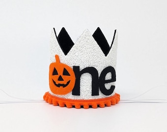 May include: White felt crown with black felt accents and an orange felt pumpkin with a jack-o'-lantern face. The crown has the word "one" in black felt letters and an orange felt pom-pom trim.