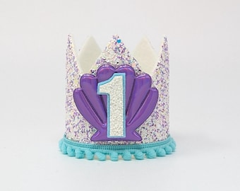 May include: A crown featuring a white and purple glitter finish with a scalloped top edge. A large, shiny purple seashell applique is centered on the front, displaying a white glittery number one. The base is trimmed with a light blue pom-pom fringe.