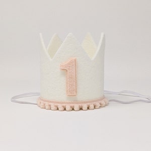 May include: White felt crown with a pink felt number one on the front. The crown has a pink pom-pom trim around the bottom.