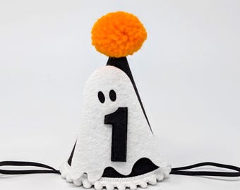 Op de afbeelding: Zwarte en witte vilten feesthoed met een spookontwerp en het nummer 1. De hoed heeft een oranje pompon bovenop en een geschulpte rand. Een zwart elastiek is bevestigd om te dragen.