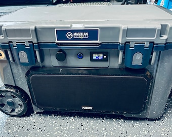 May include: A gray Magellan Outdoors Pro cooler with a built-in Fusion speaker. The cooler has a digital display and a blue light. The cooler is on wheels and has a handle.