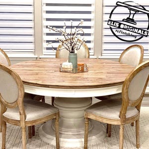 Puede incluir: Una mesa de comedor redonda de madera con una base blanca y cuatro sillas. La mesa tiene una tapa de madera clara y una base pintada de blanco. Las sillas son de madera con un acabado marrón claro y tienen un asiento de tela blanco. Un pequeño ramo de flores blancas está en un jarrón azul sobre la mesa.
