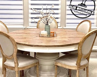 May include: A round wooden dining table with a white base and four chairs. The table has a light wood top and a white painted base. The chairs are made of wood with a light brown finish and have a white fabric seat. A small bouquet of white flowers is in a blue vase on the table.