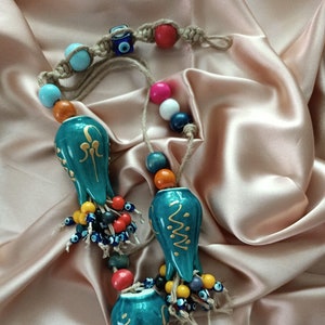 Op de afbeelding: Een turquoise blauwe keramische ornament met gouden details en een kwast van kleurrijke kralen en blauwe boze oog charmes. De ornament hangt aan een touwtje van kralen en touw.