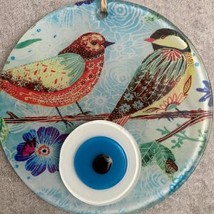 May include: A round glass wall hanging featuring two colorful birds perched on a branch. The background is light blue with floral patterns. A blue eye-shaped bead hangs above the glass disc, adding a decorative touch.