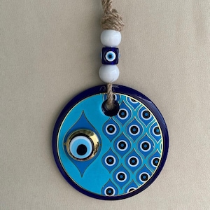 Könnte beinhalten: Ein blauer und weißer Keramik-Schutzamulet-Wandbehang mit goldener Akzent. Der Behang zeigt ein blaues Schutzamulet mit einer weißen Pupille und einem goldenen Rand. Das Schutzamulet ist von einem blauen und weißen Muster aus Kreisen und Ovalen umgeben. Der Behang ist an einer Schnur mit weißen und blauen Perlen befestigt.