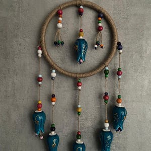 May include: A handcrafted dream catcher with a jute-wrapped hoop and hanging teal ceramic tulip-shaped ornaments. Colorful wooden beads in red, white, and blue are strung along the hanging cords, adding a decorative touch to this wall hanging.