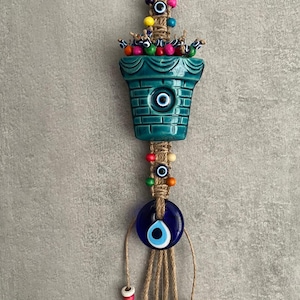 May include: A turquoise ceramic well-shaped wall hanging with a blue eye design. The hanging features a macrame design with colourful beads and a large blue eye charm. The well has a small eye design in the centre.