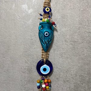 Könnte beinhalten: Eine blaue Keramikvase mit einem blauen bösen Blick-Anhänger. Die Vase ist mit weißen Blumenmustern verziert. Die Vase hängt an einer Schnur aus Bindfaden mit blauen und weißen bösen Blick-Anhängern und bunten Perlen.