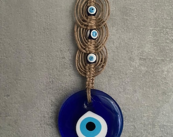 May include: A decorative wall hanging featuring a large, round, deep blue glass eye with a white and light blue iris. The eye is suspended from a series of three woven, circular, tan-colored macrame rings, each with a smaller blue eye. A unique home decor item.