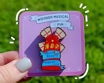 May include: Wooden musical pin with a red and white Ferris wheel design. The pin has a blue ribbon with the words "Truth, Beauty, Freedom, Love" written on it. The pin is on a pink card with the words "Wooden Musical Pin" written on it.