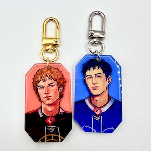May include: Two acrylic keychains with portraits of men. One keychain has a gold clasp and a red background, the other has a silver clasp and a blue background. Both keychains have a gold-colored metal ring.