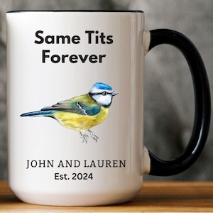 White ceramic mug with a black handle featuring a humorous bird-themed design. The mug displays an illustration of a blue tit bird with the phrase 'Same Tits Forever.' Personalized with 'John and Lauren' and 'Est. 2024.' Cozy setting with a soft blanket in the background.