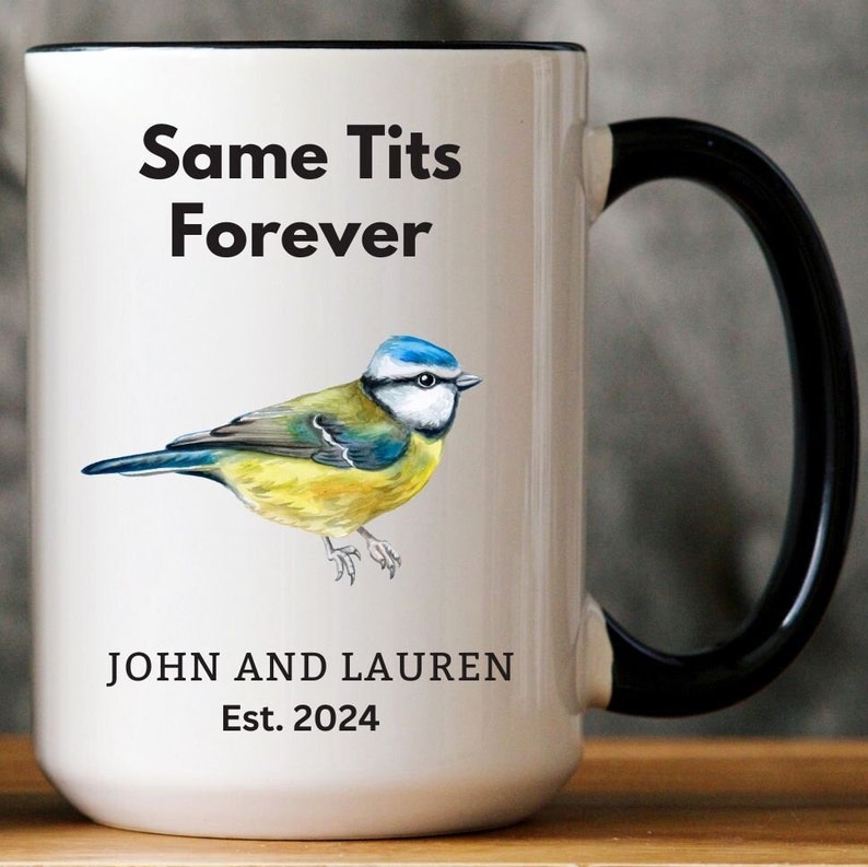 White ceramic mug with a black handle featuring a humorous bird-themed design. The mug displays an illustration of a blue tit bird with the phrase 'Same Tits Forever.' Personalized with 'John and Lauren' and 'Est. 2024.' Cozy setting with a soft blanket in the background.