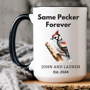 White ceramic mug with a black handle featuring a humorous bird-themed design. The mug displays a woodpecker illustration with the phrase 'Same Pecker Forever.' Personalized with 'John and Lauren' and 'Est. 2024.' Cozy setting with a soft blanket in the background.
