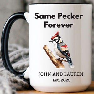 White ceramic mug with a black handle featuring a humorous bird-themed design. The mug displays a woodpecker illustration with the phrase 'Same Pecker Forever.' Personalized with 'John and Lauren' and 'Est. 2025.' Cozy setting with a soft blanket in the background.