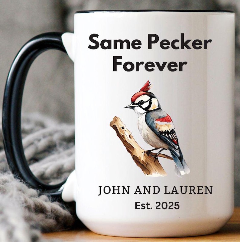 White ceramic mug with a black handle featuring a humorous bird-themed design. The mug displays a woodpecker illustration with the phrase 'Same Pecker Forever.' Personalized with 'John and Lauren' and 'Est. 2025.' Cozy setting with a soft blanket in the background.
