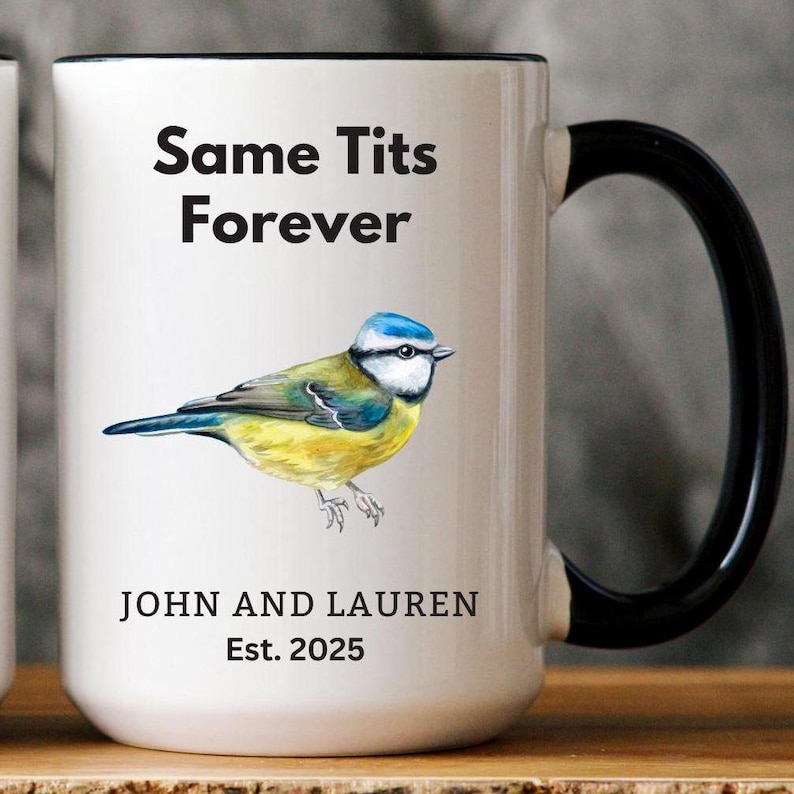 White ceramic mug with a black handle featuring a humorous bird-themed design. The mug displays an illustration of a blue tit bird with the phrase 'Same Tits Forever.' Personalized with 'John and Lauren' and 'Est. 2025.' Cozy setting with a soft blanket in the background.