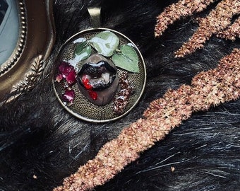May include: A round pendant necklace with a resin-filled image of a pair of red lips with fangs, surrounded by green leaves and red berries. The pendant is set in a bronze-colored metal frame.