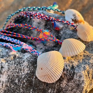 Peut inclure: Cordons tressés multicolores avec des coquillages attachés. Les cordons sont rose, bleu, blanc, orange et jaune. Les coquillages sont blancs et ont une texture naturelle.