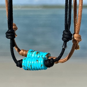May include: Two cords, one brown and one black, are tied together with a turquoise blue bead. The cords are hanging against a blurred background of a beach and ocean.