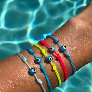 May include: A stack of colourful string bracelets with blue evil eye charms. The bracelets are worn on a person's wrist against a blue water background.