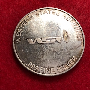 May include: A silver coin with the text "WESTERN STATES REFINING" around the top edge. The centre features the letters "WSR" and a flame symbol. The bottom edge reads ".999 FINE SILVER".