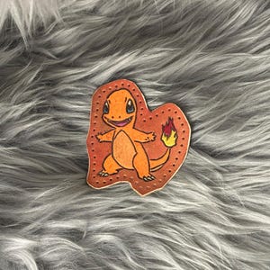 May include: An orange Charmander character illustration on a brown, heart-shaped leather patch. The patch has a stitched border and is set against a gray, furry background. The Charmander has a flame on its tail.