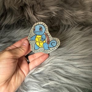 Puede incluir: Un personaje de Squirtle Pokemon pintado a mano sobre un fondo azul claro con detalles marrones y amarillos. El personaje está rodeado por un borde punteado.