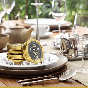 Puede incluir: Una pila de tres latas doradas de caviar en un plato blanco con cubitos de hielo. La lata superior tiene una etiqueta negra con letras doradas que dice "The World's Caviar Malossol".