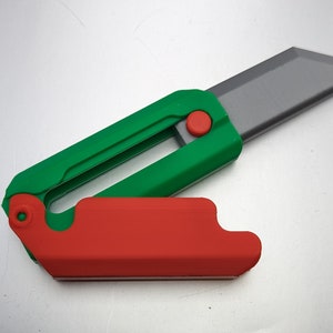 Puede incluir: Un cuchillo de utilidad plegable verde y rojo con una hoja gris. El cuchillo tiene un botón rojo en el lateral.