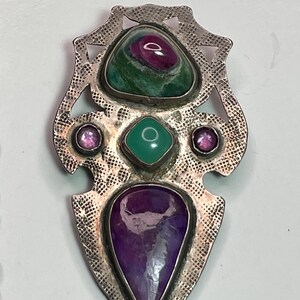 May include: A silver pendant with an ornate design, featuring a large, teardrop-shaped purple stone at the bottom, a square green stone in the center, and a ruby-colored stone at the top. Two smaller pink stones flank the green stone.