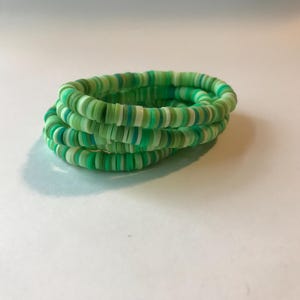 Puede incluir: Cuatro pulseras apiladas hechas de pequeñas cuentas redondas y planas en tonos verdes, blancos y azules. Las pulseras están enrolladas juntas, creando un efecto en capas. Las cuentas parecen estar hechas de arcilla polimérica o material similar.