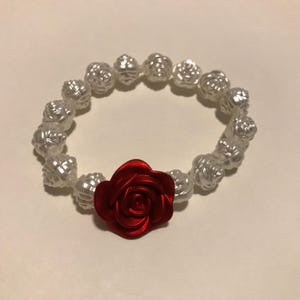 May include: A white beaded bracelet with a central red rose. The bracelet is made up of white, rose-shaped beads. A bright red rose is the focal point, providing a striking contrast. This bracelet is a fashionable accessory, suitable for various occasions.