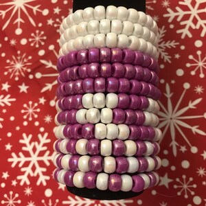 May include: A stack of beaded bracelets in alternating rows of white and iridescent purple beads. The bracelets are stacked on a black base, with a red background featuring white snowflakes.