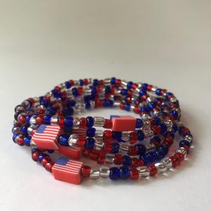 Puede incluir: Un conjunto de pulseras de cuentas rojas, blancas y azules, con pequeños dijes de la bandera estadounidense. Las pulseras están hechas de pequeñas cuentas redondas y están dispuestas en espiral. La combinación de colores es patriótica.